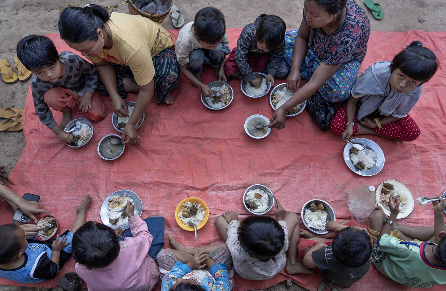 Several children sit on a blanket on the ground eating from bowls